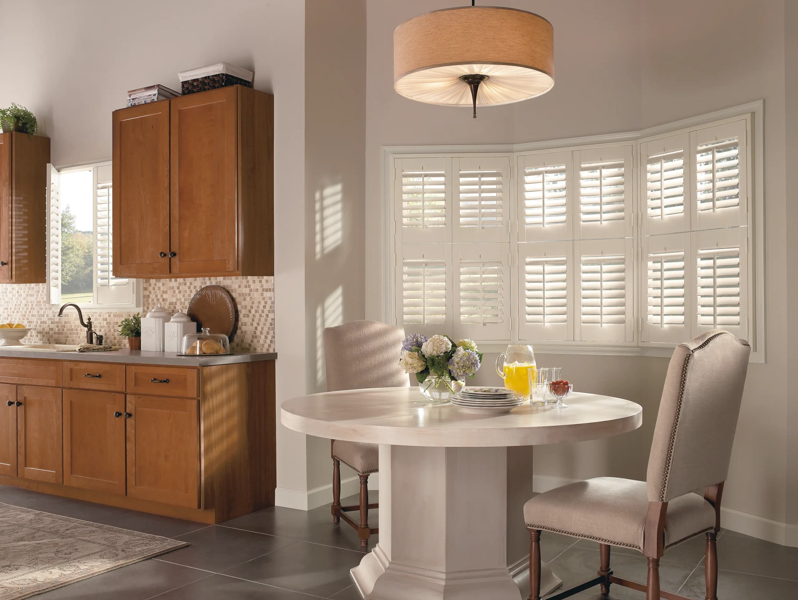 A room corner with white plantation shutters in the foreground and a roman shade on the adjacent window.