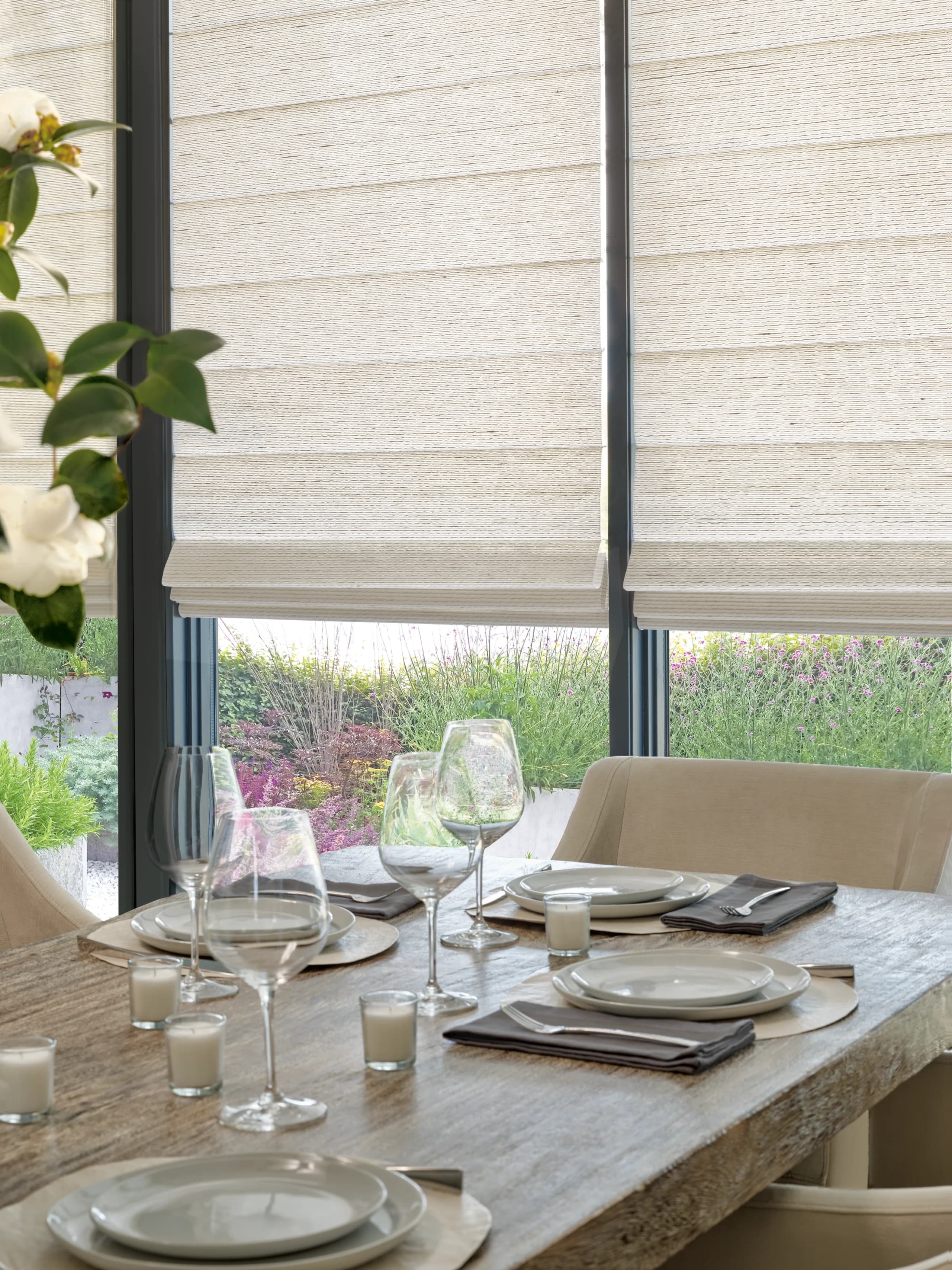 The Alustra® Collection of Woven Textures® Roman Shades in a cream weave, lowered over dining room doors above a table set for dinner, with a garden beyond the glass.