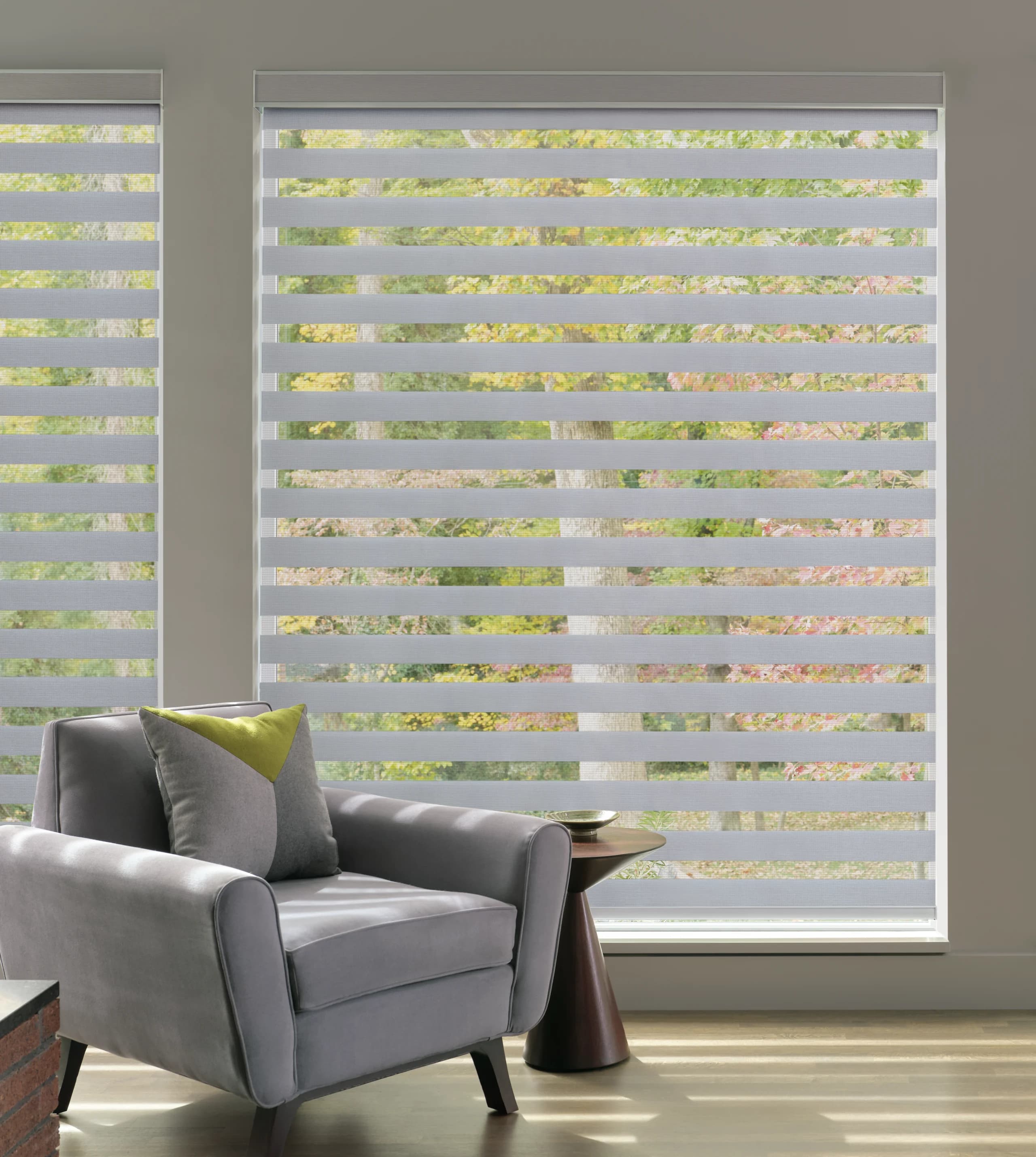 A close view of a gray Designer Banded Shade with the bands aligned on autumn foliage, beside a gray armchair and a dark wood side table.