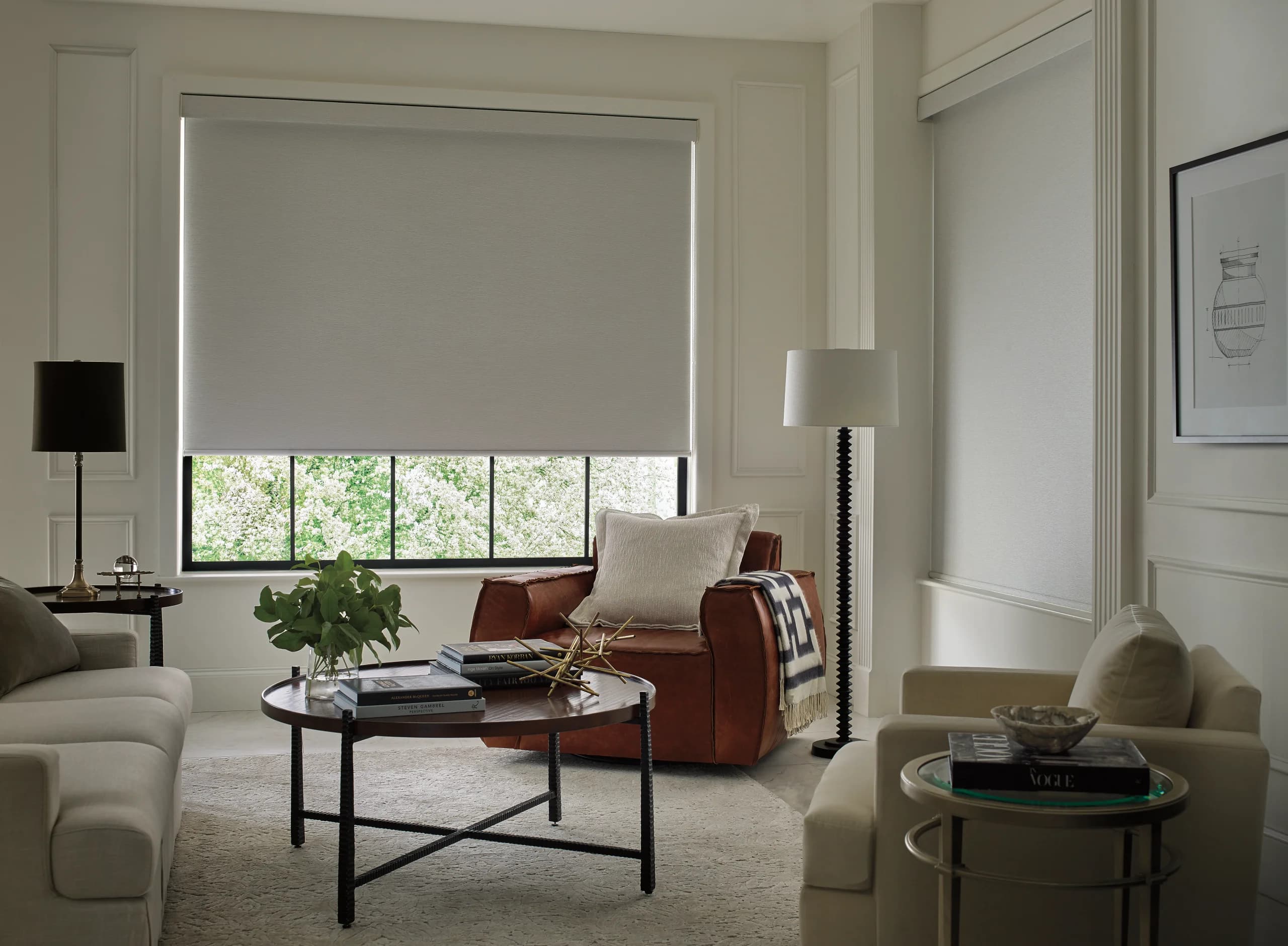 A cream Designer Roller Shade over a black-framed picture window in a formal living room, with flowering trees outside and a leather armchair beside it.