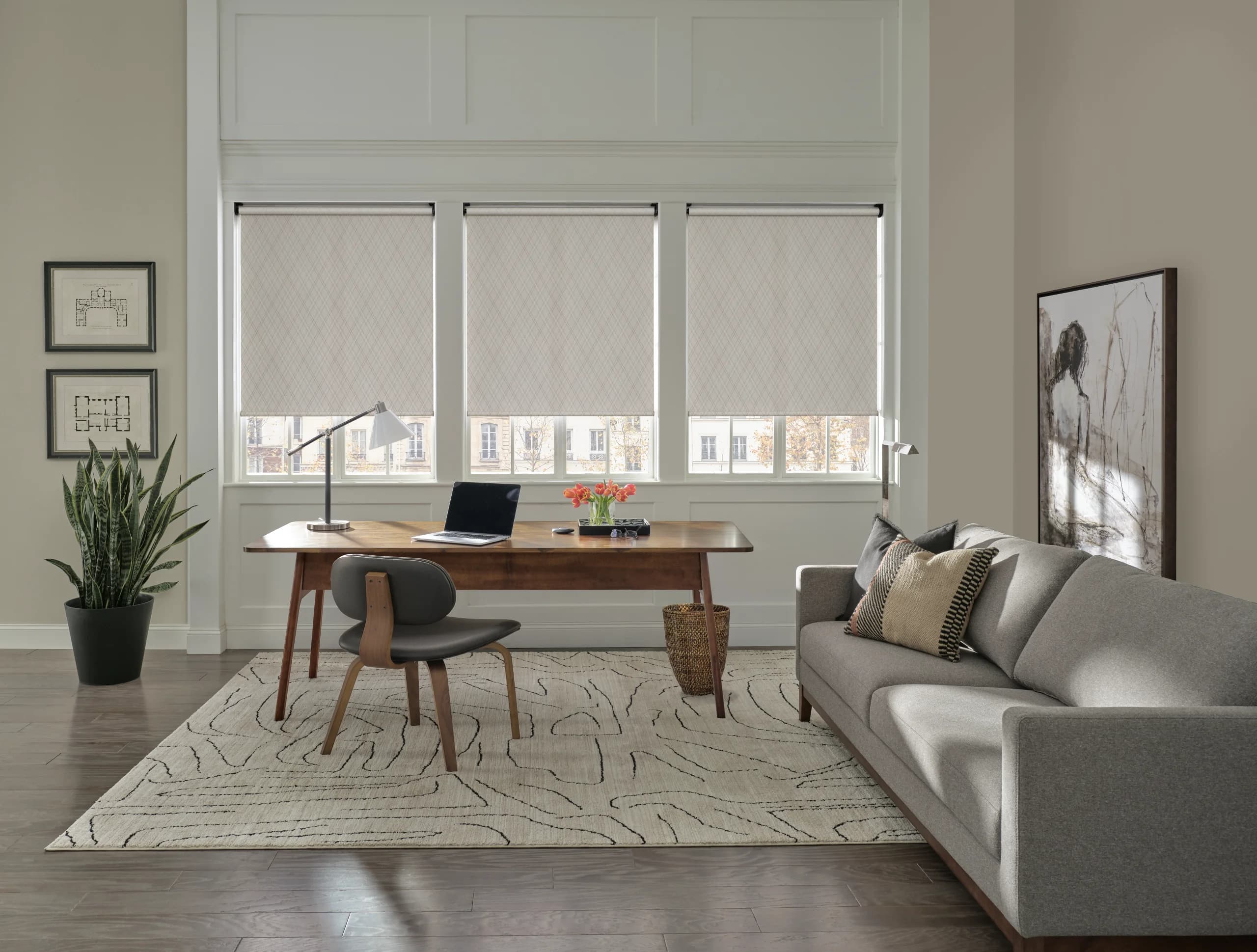 A Designer Roller Shade lowered behind a desk in a living-room office, filtering the daylight across the work surface.