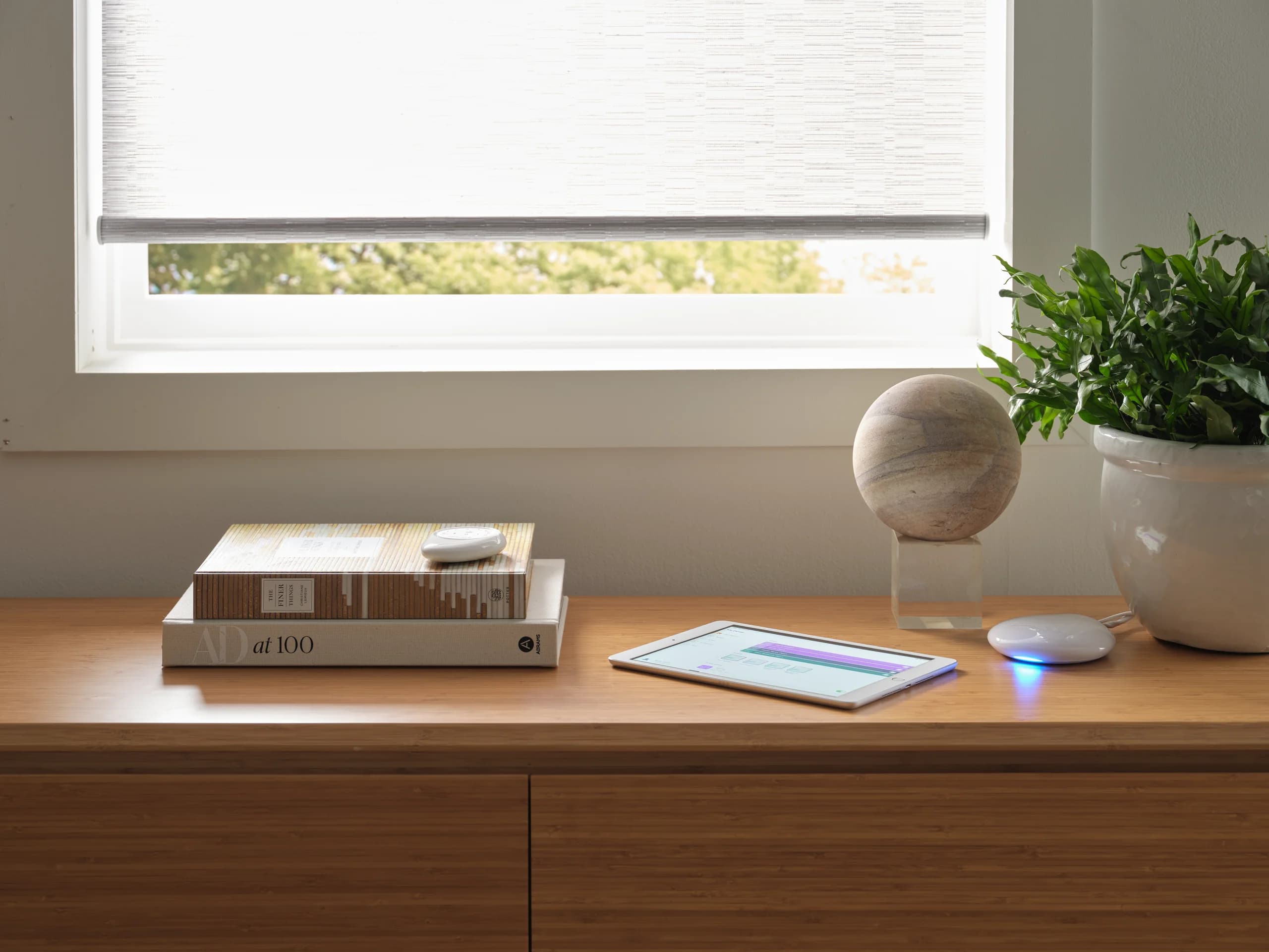 A soft white Designer Roller Shade lowered over a sunlit window, with a slim remote resting on a stack of books on the wood credenza below.