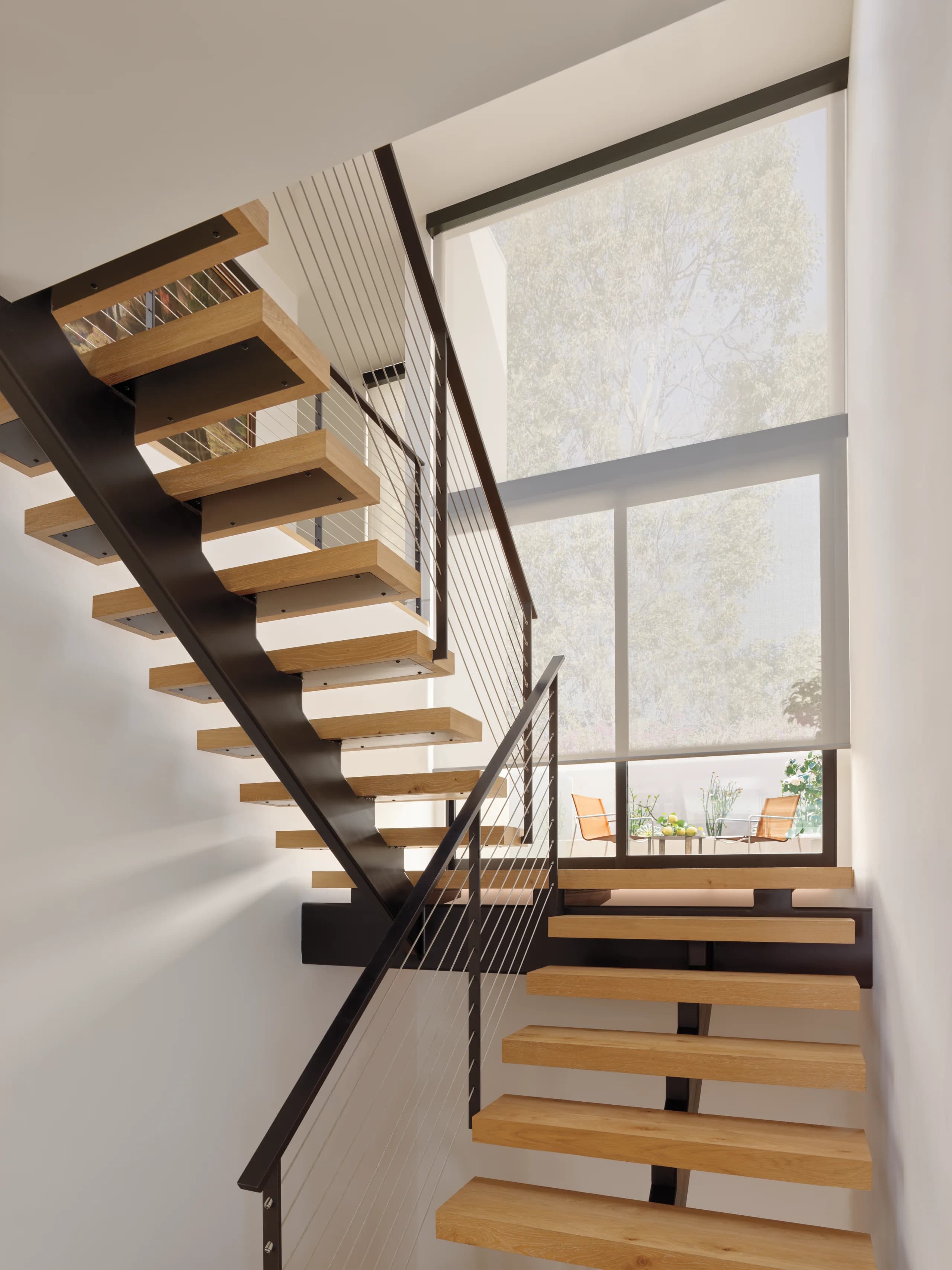 A Designer Screen Shade on a tall stairway window, keeping the trees and patio in view while cutting the glare on the wood treads.