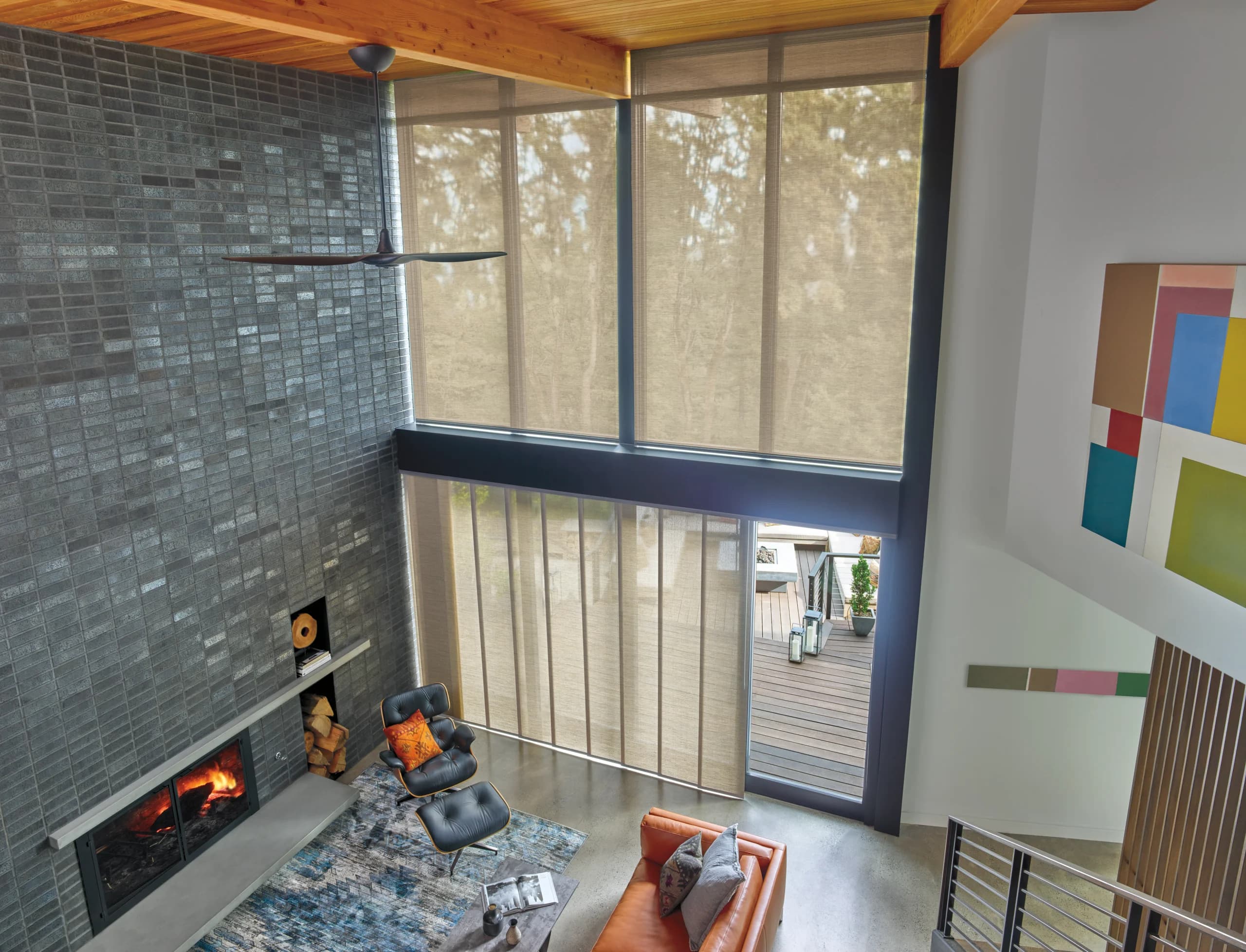 Designer Screen Shades covering a two-story window wall in a living room, with trees showing through the woven fabric above a brick fireplace.