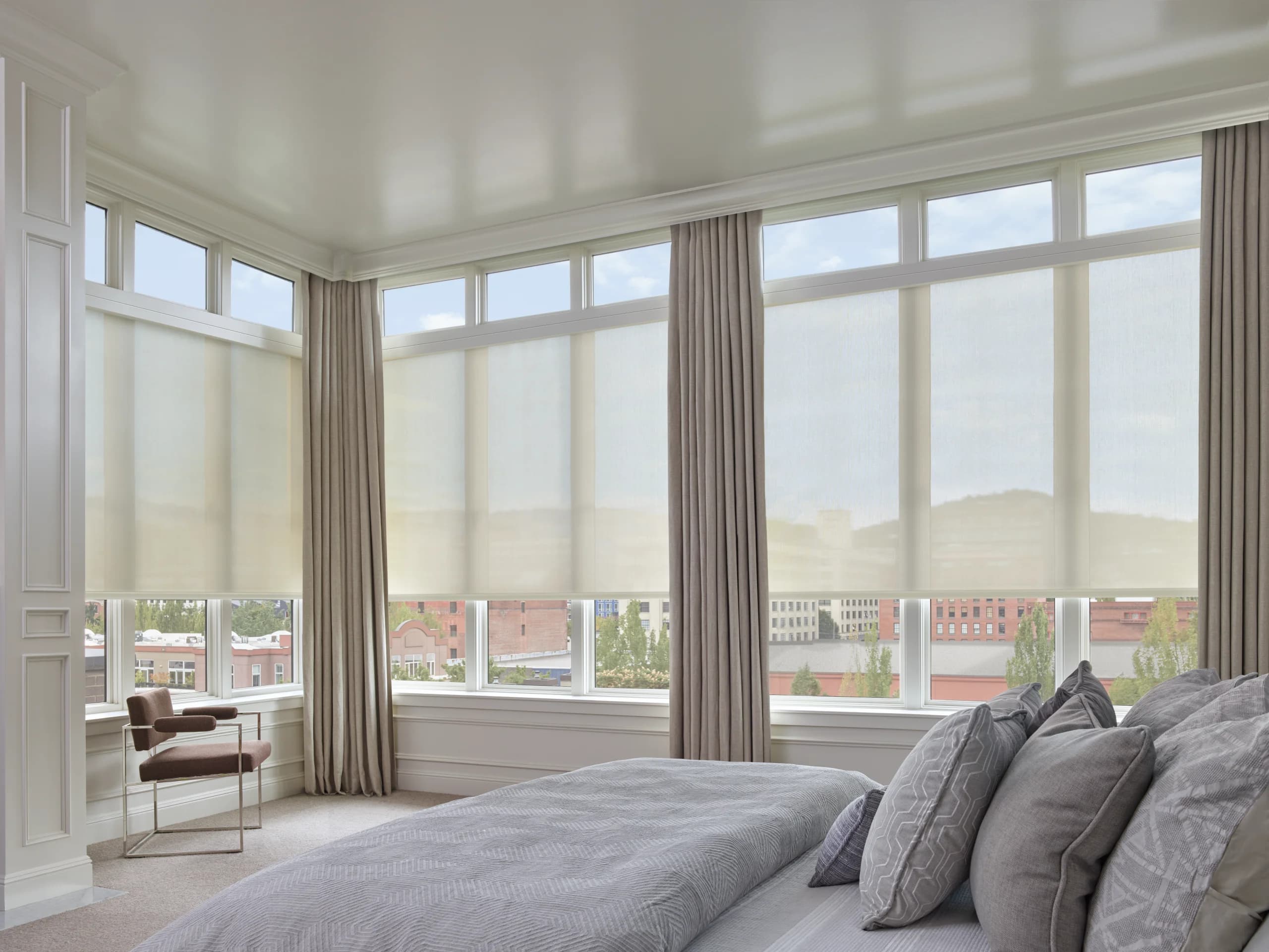 Designer Screen Shades lowered partway across the wraparound windows of a corner bedroom, layered under floor-length drapery panels.