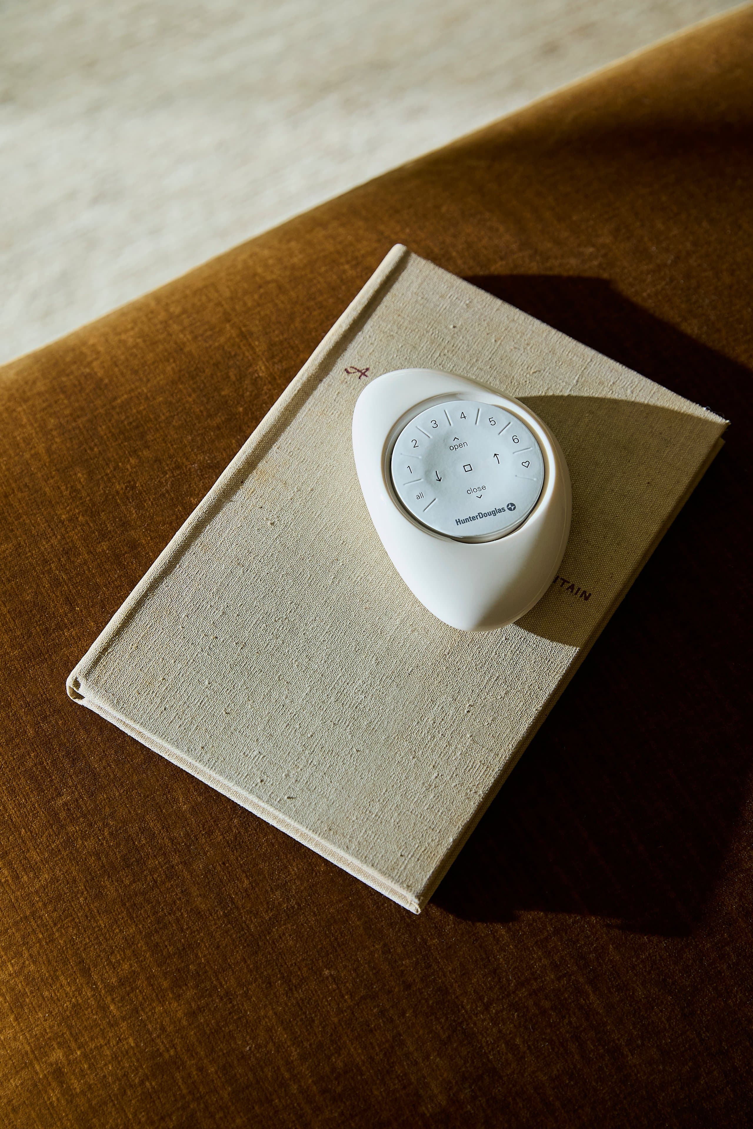 A rounded white PowerView® Automation remote with numbered channel buttons and open and close controls, resting on a linen-bound book on gold velvet.