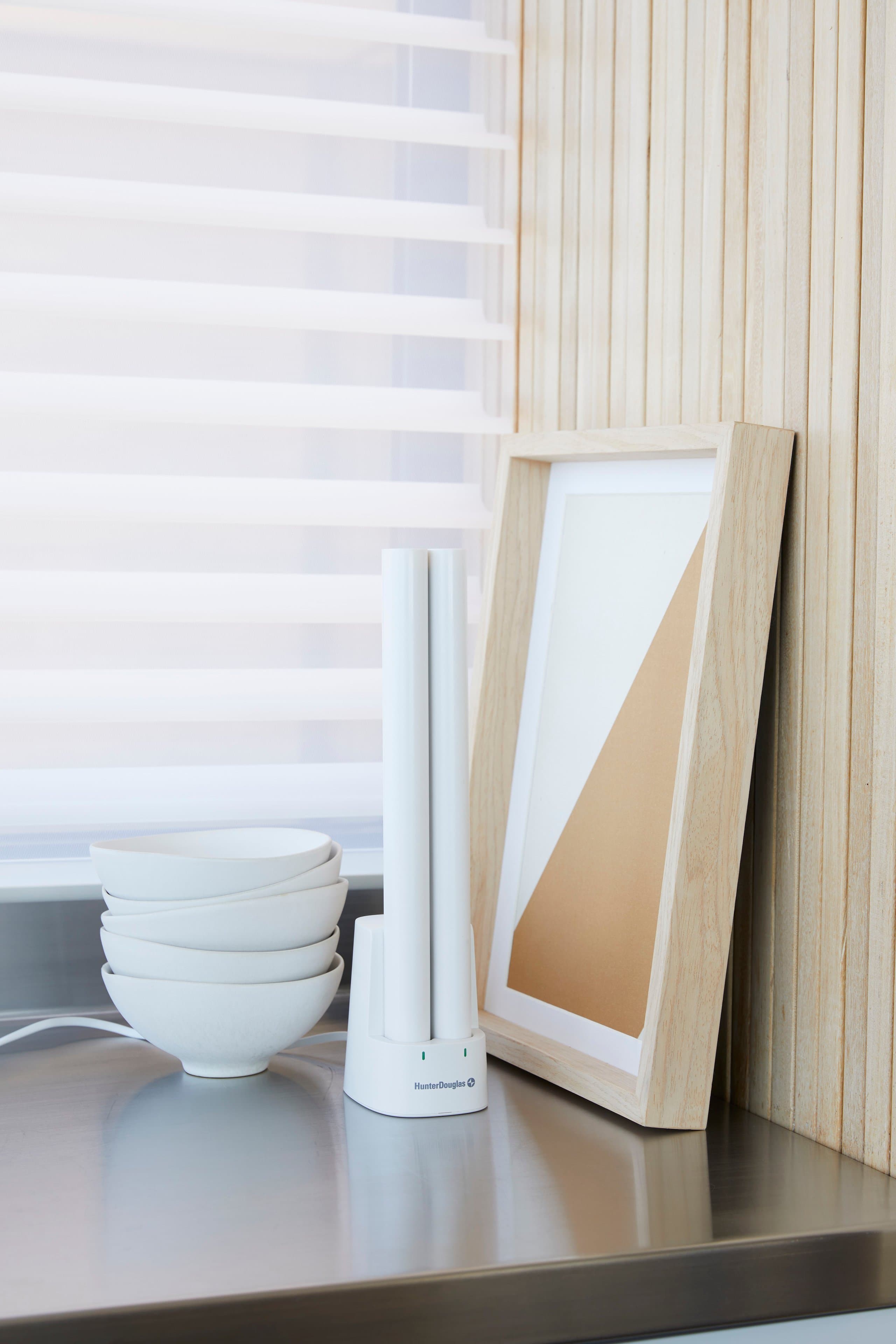 A rechargeable PowerView® Automation battery wand standing upright in its charging base on a kitchen counter, in front of a lowered shade.