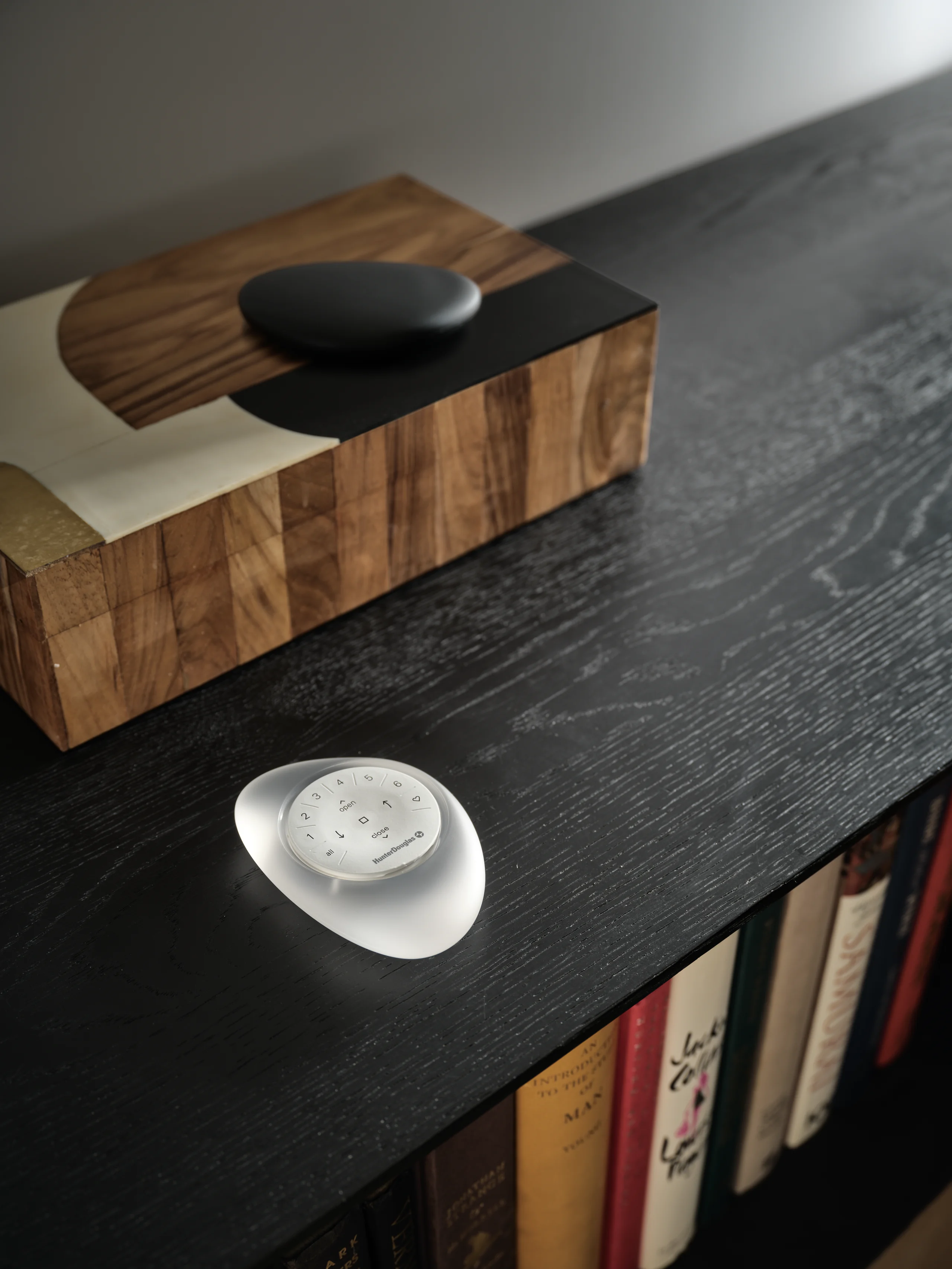 A frosted PowerView® Automation remote on a black oak console beside a walnut box and a row of books.