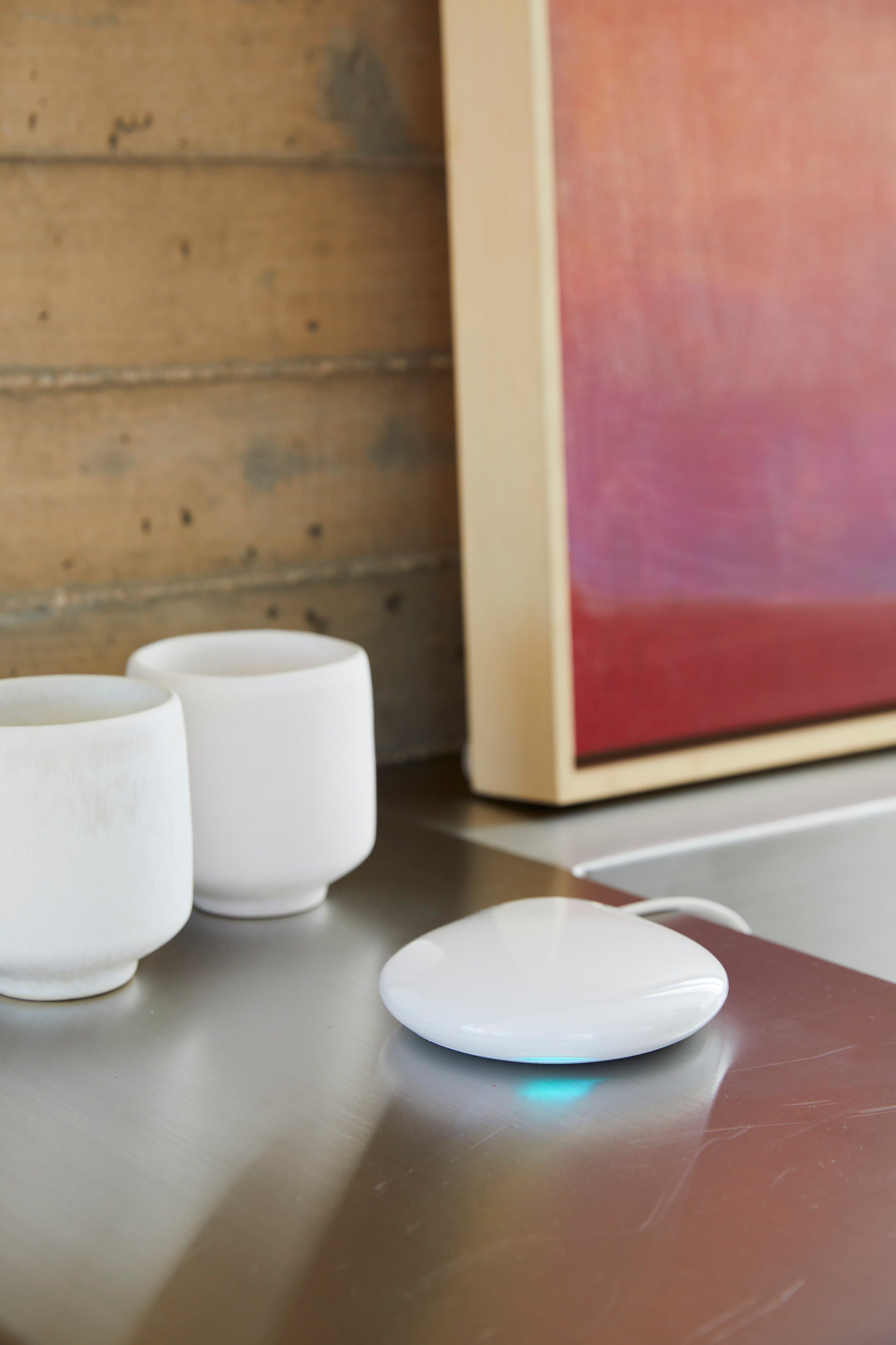 A compact white PowerView® Automation gateway glowing softly on a stainless steel counter beside two white ceramic cups.