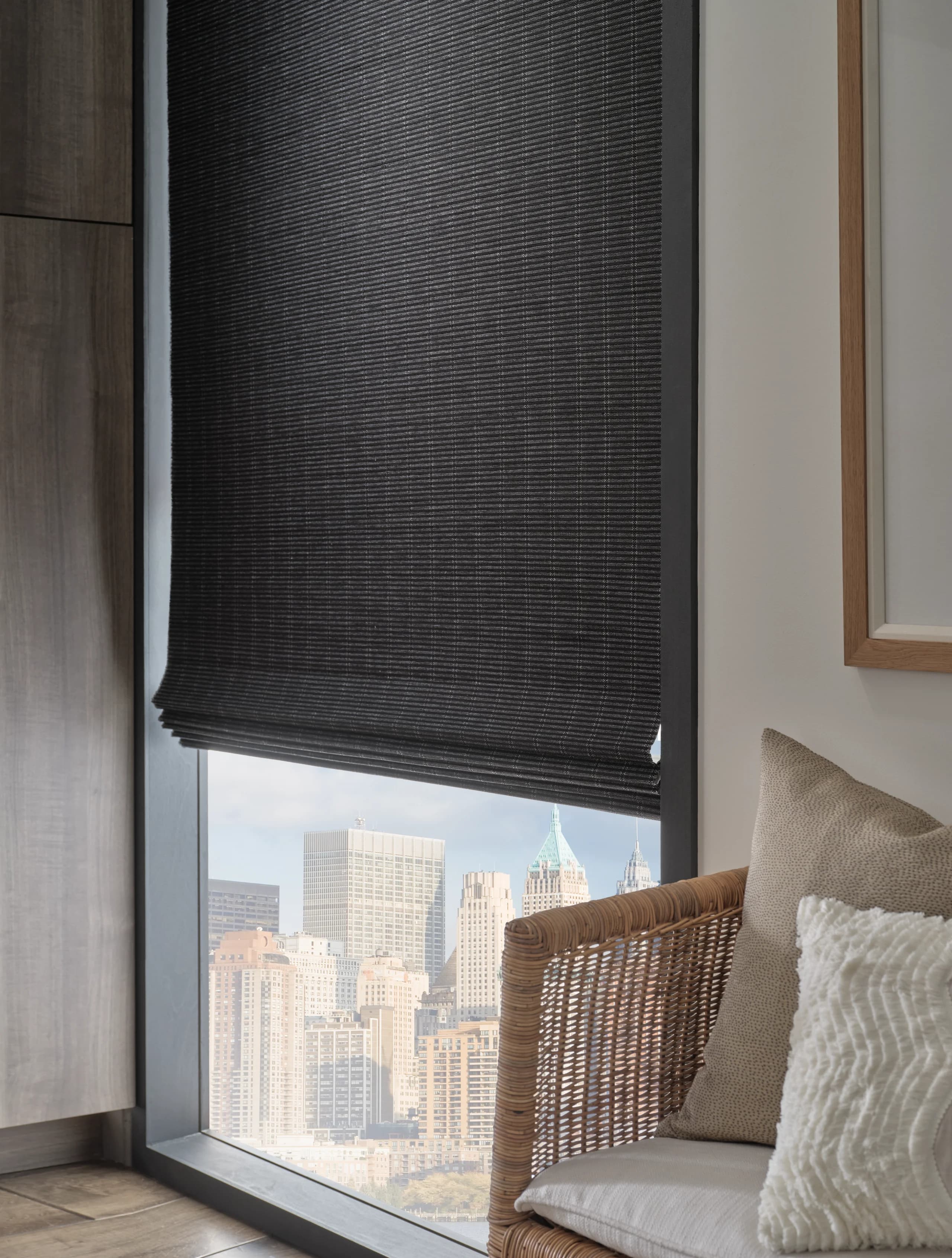 A close view of the folds and weave of a black Provenance® Woven Wood Shade raised above a city skyline.