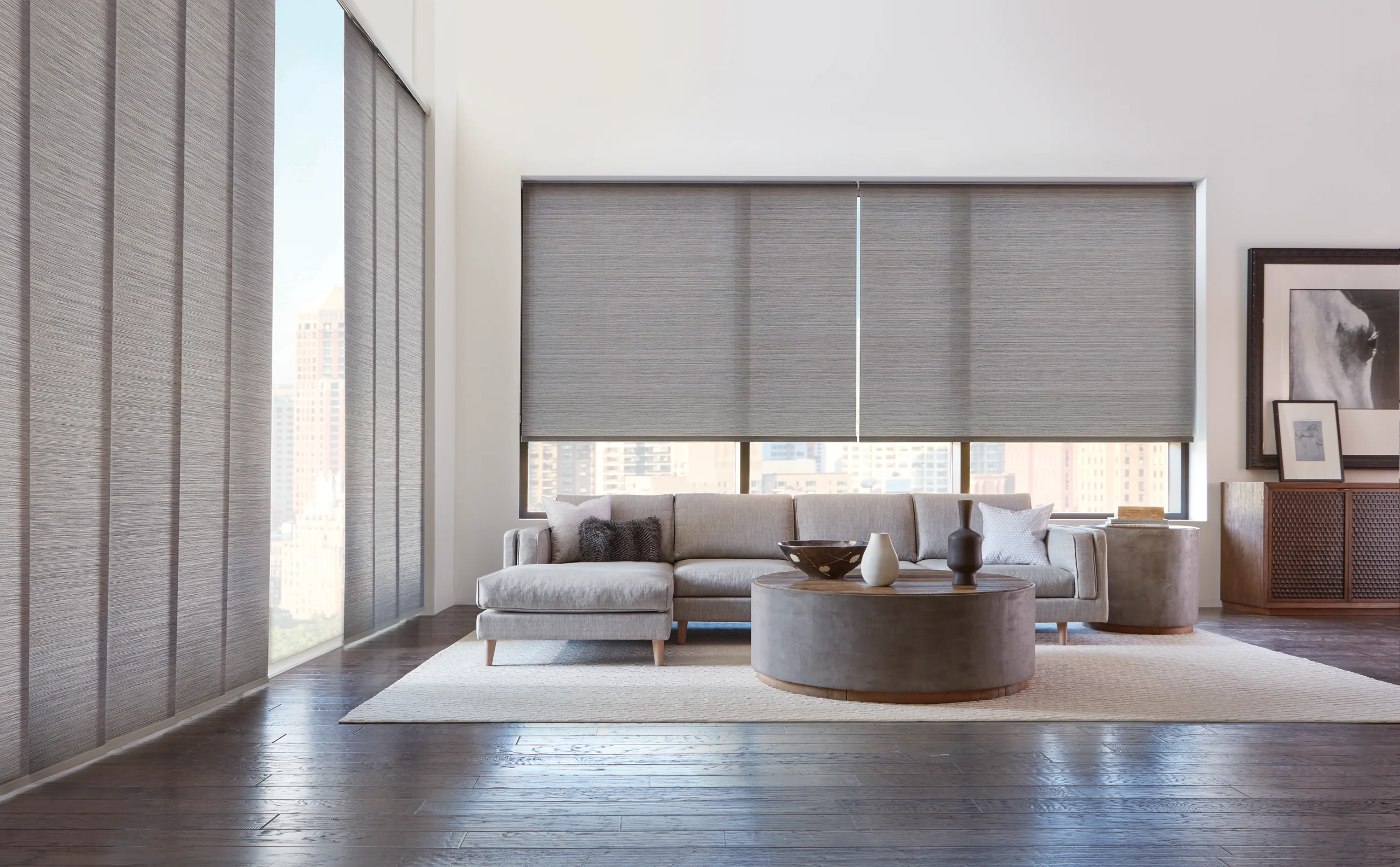 Designer Roller Shades in a woven gray fabric lowered across a wide living room window, with a city skyline showing beneath the hem.