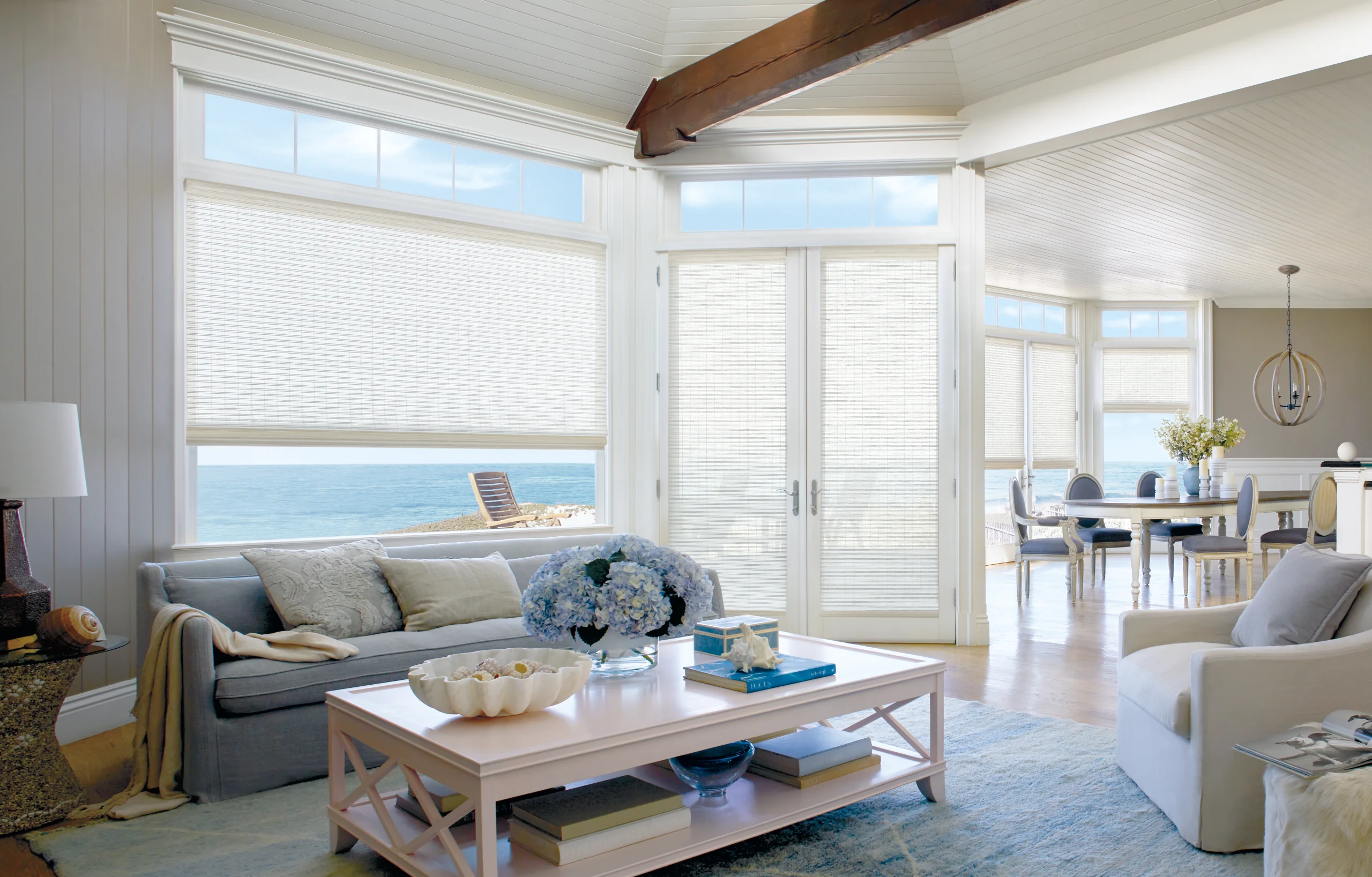 Provenance® Woven Wood Shades in a pale weave lowered across an ocean-view window and the French doors of a coastal living room.